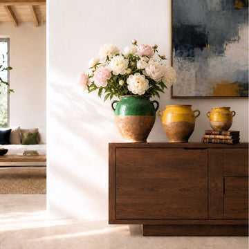 Three antique French confit pots with green and yellow glaze on a modern sideboard, one filled with pale peonies.