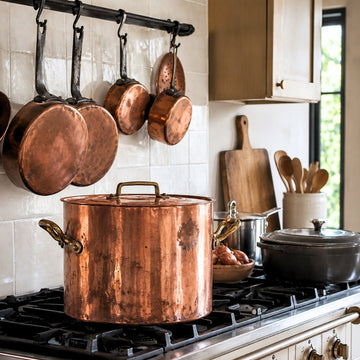 Antique French copper stockpot on a kitchen range beneath hanging copper pans and a colander in an elegant modern kitchen.