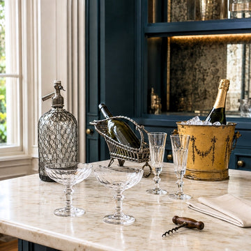 Antique French drinkware with crystal coupes and flutes, a soda siphon, wine cradle and champagne bucket on marble.