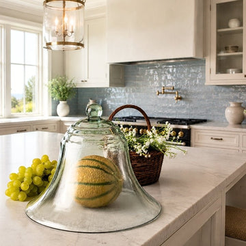 Antique French glass garden cloche covering a melon on a marble island in an elegant coastal kitchen.