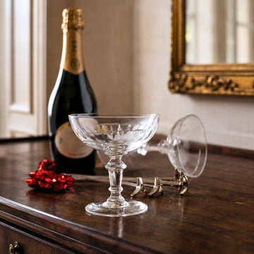 Antique French cut-crystal champagne coupes beside a champagne bottle and cork on a polished wooden sideboard.