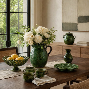 Green-glazed antique Provençal pottery, including a flower-filled pitcher, cups, bowls and jars, arranged on a dining table.