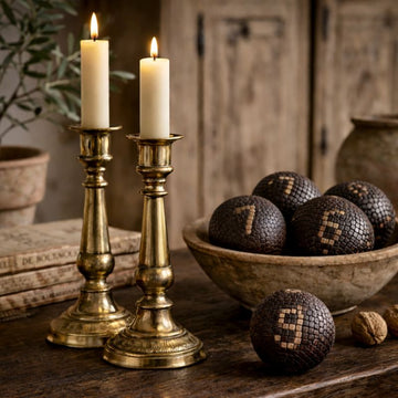 Two lit brass candlesticks with white candles sit on a wooden table beside a bowl filled with numbered textured balls, with rustic decor and books in the background.