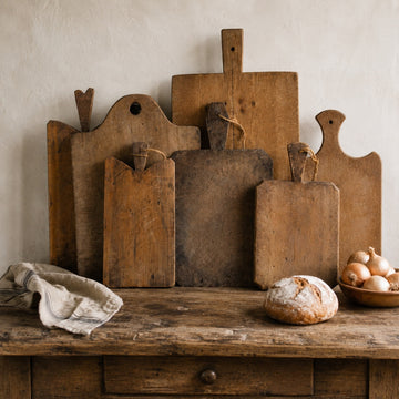 Collection of antique wooden cutting boards with worn patina, displayed on a rustic table with linen, bread, and onions.