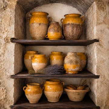 Three wooden shelves in a stone alcove display rustic clay pots with yellow glaze, a bowl of baguettes, and bundles of dried lavender. The scene has a vintage, Mediterranean feel.
