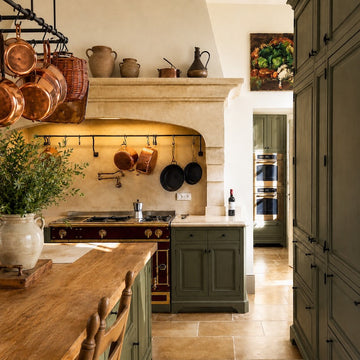 A rustic kitchen with green cabinets, a wooden island, copper pots hanging above a range, and decorative pottery on a stone mantel. Warm lighting and a still-life painting create a cozy atmosphere.