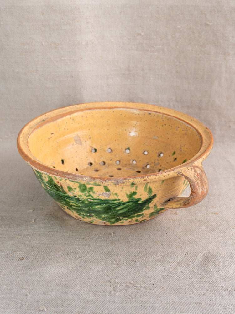 Antique French jaspe-glazed earthenware colander with yellow and green finish, single handle, on beige textured fabric.