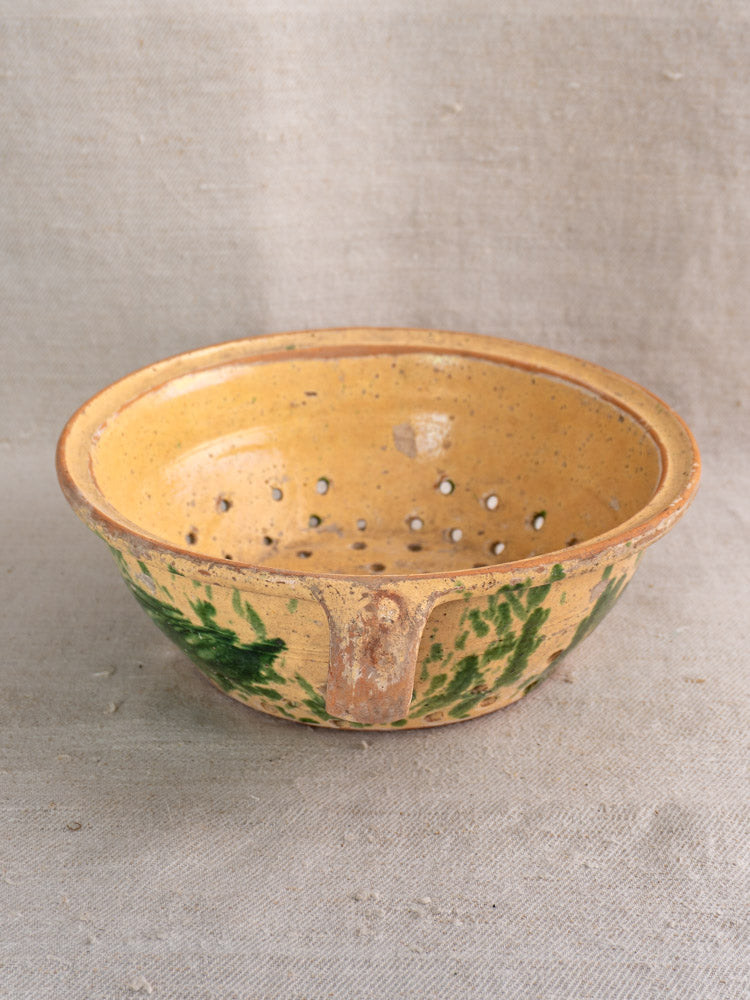 Antique French earthenware colander with jaspe yellow glaze and green leaf pattern on beige fabric background.