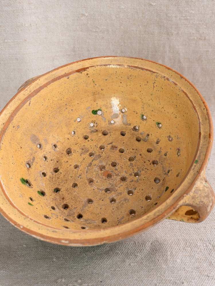 Antique French earthenware colander with jaspe glaze, yellow-brown finish, green spots, on textured fabric.