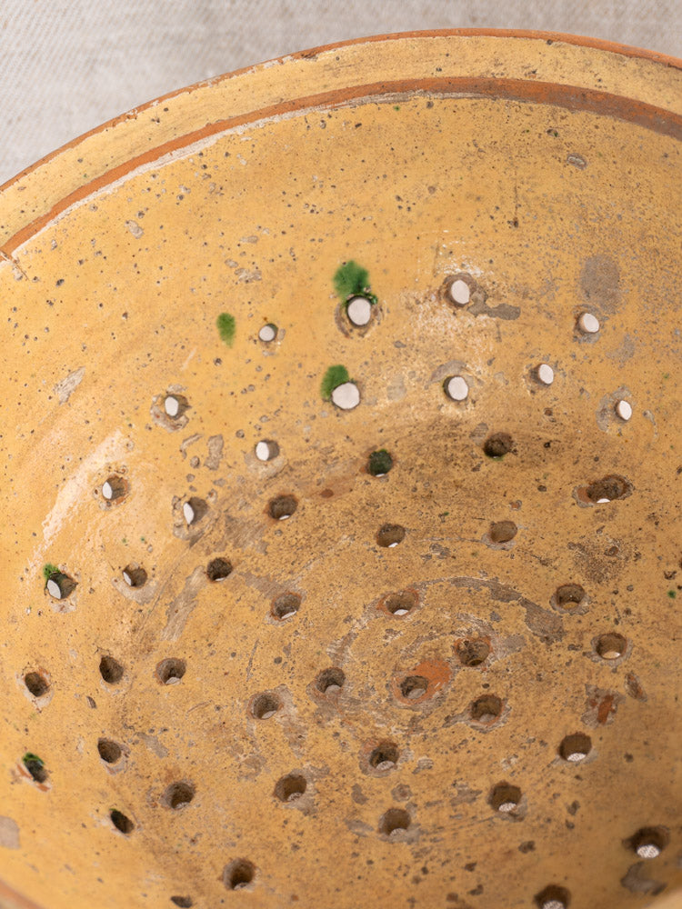 Antique French earthenware colander, late 19th century, jaspe glaze in yellow-brown with green spots, on neutral fabric background.