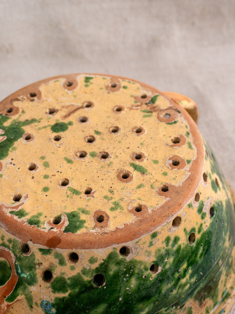 Antique French earthenware colander base with green-yellow jaspe glaze and drainage holes on textured neutral fabric.