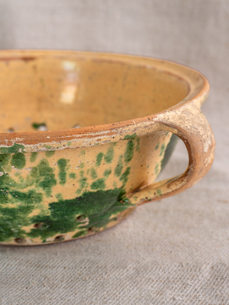 Antique French earthenware colander with jaspe glaze, rustic yellowish-brown handle, green speckled finish on beige surface.