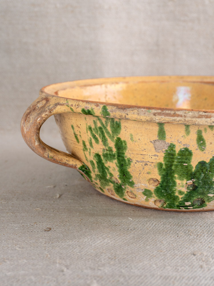 Antique French earthenware colander with jaspe glaze, rustic yellow and green, on neutral fabric background.
