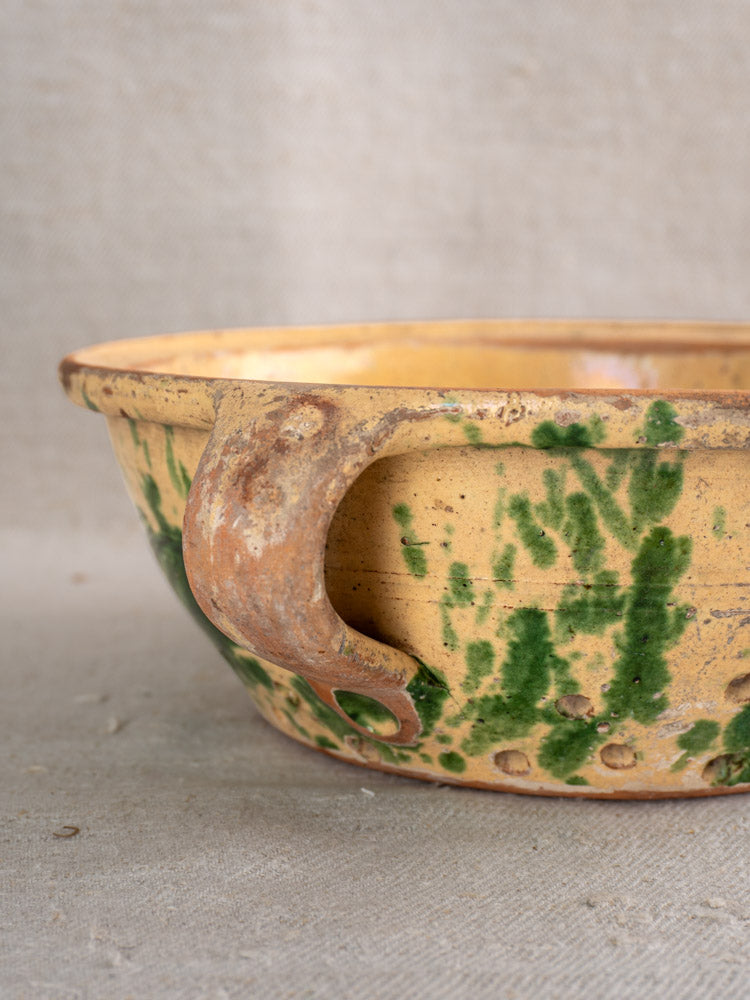 Antique French earthenware colander with yellow and green jaspe glaze, rusted handle, on neutral fabric background.