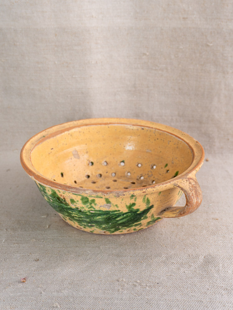 Antique French earthenware colander with jaspe glaze, green accents, and one handle on textured beige fabric.