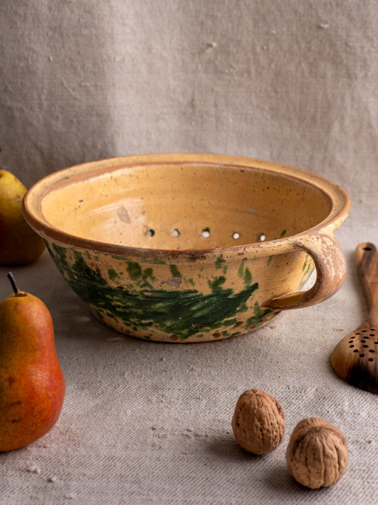 Antique French earthenware colander with jaspe glaze, green accents, and one handle on textured beige fabric.