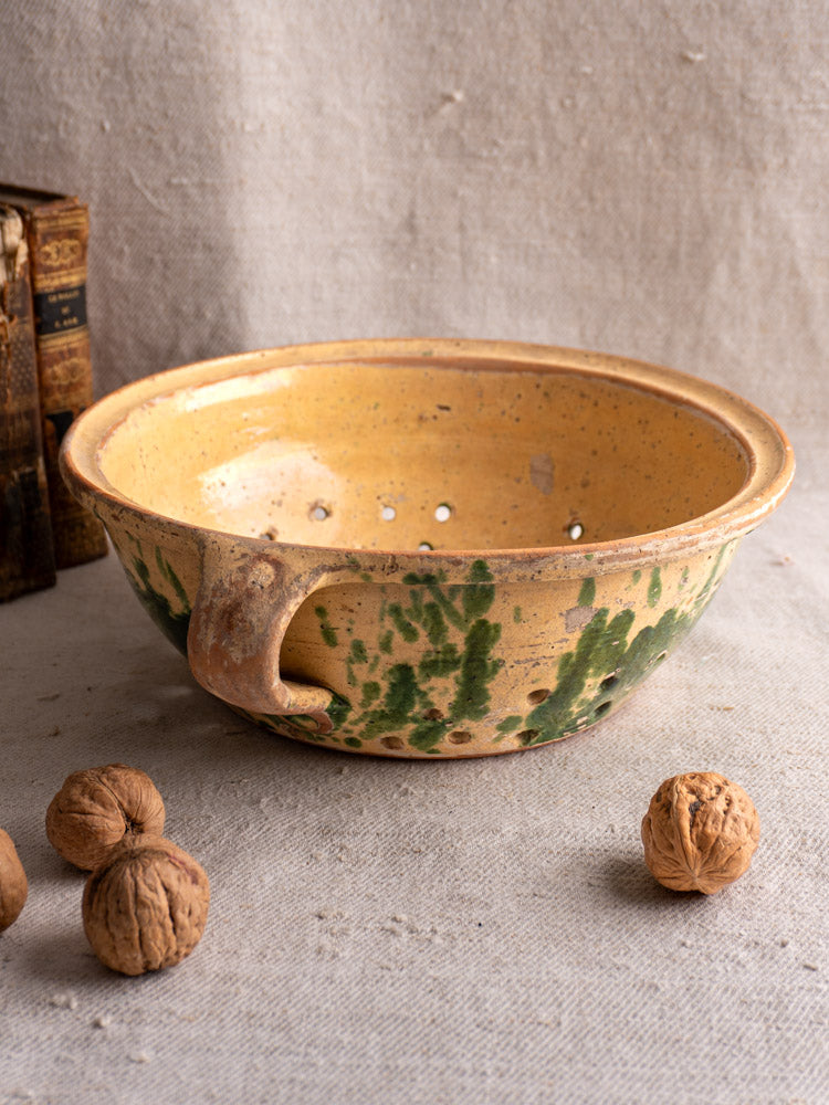 Antique French earthenware colander with yellow jaspe glaze and green accents, displayed with walnuts and vintage books.