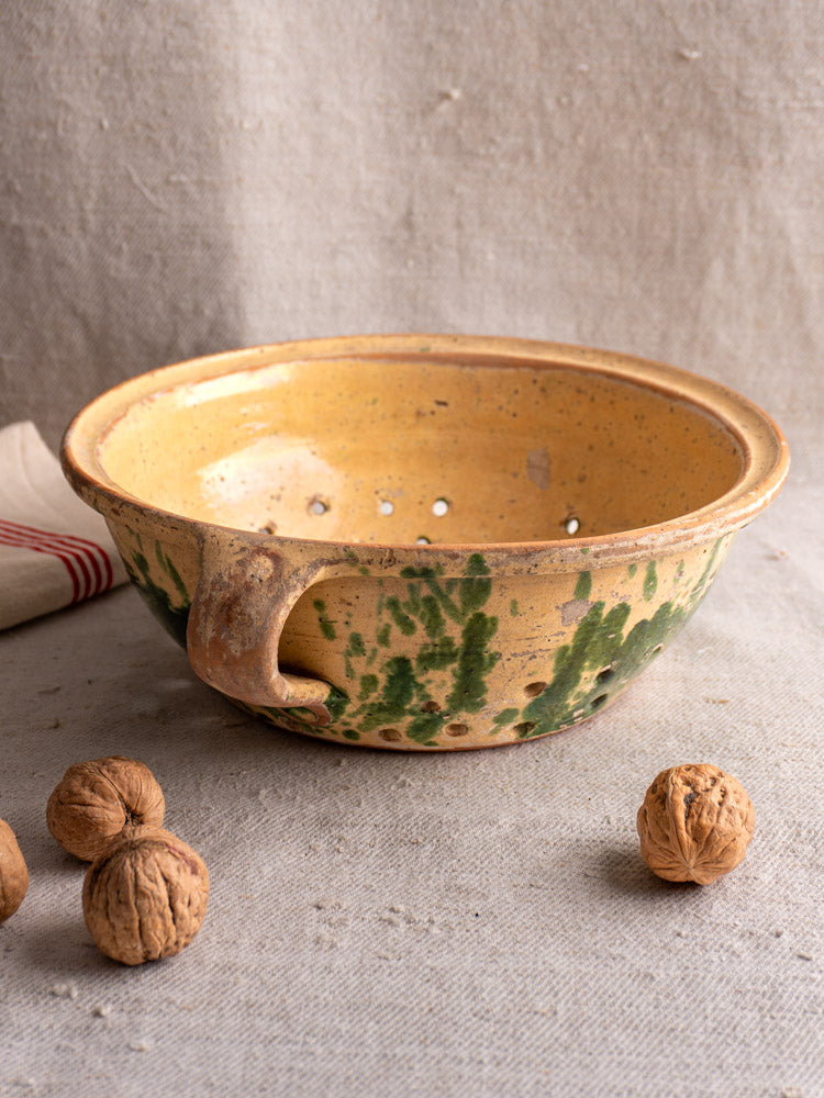 Antique French earthenware colander with jaspe glaze and handle on textured cloth, unshelled walnuts nearby, striped fabric behind.