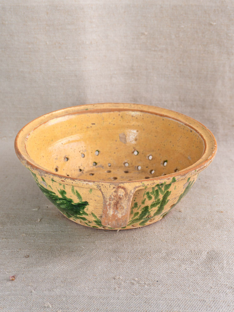 Antique French earthenware colander with jaspe glaze and green leaf motifs on a beige fabric surface.