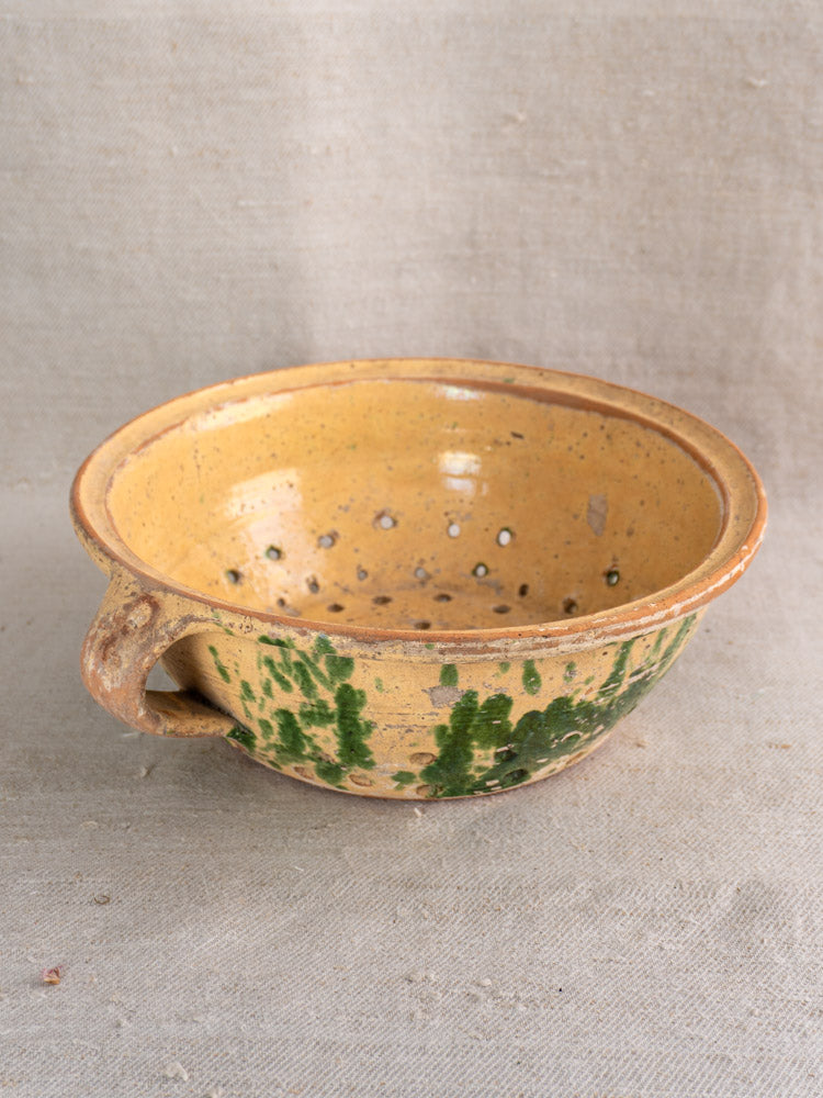Antique French earthenware colander with jaspe yellow glaze, green drips, and side holes on neutral fabric background.