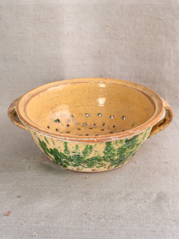 Late 19th-century French jaspe earthenware colander with two handles, yellow-green patterns, on textured beige fabric.