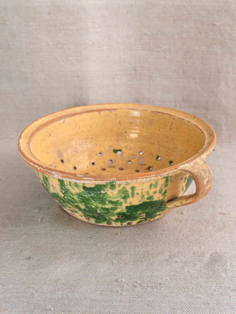 Antique French earthenware colander with jaspe glaze in yellow-brown and green, handle and base holes; neutral fabric backdrop.