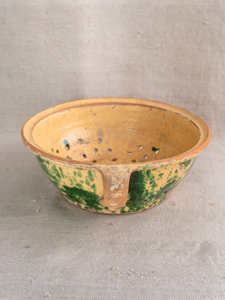 Antique French earthenware colander with yellow jaspe glaze and green spots on textured fabric surface.