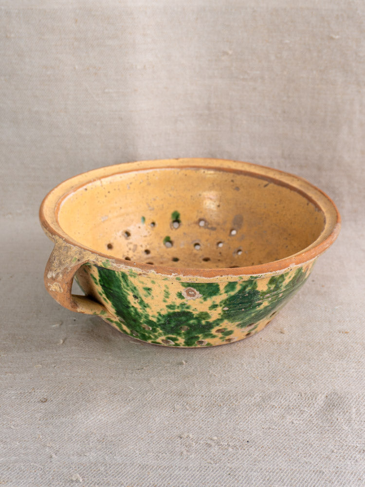 Antique French earthenware colander with yellow and green jaspe glaze, handle, and drainage holes on textured fabric background.