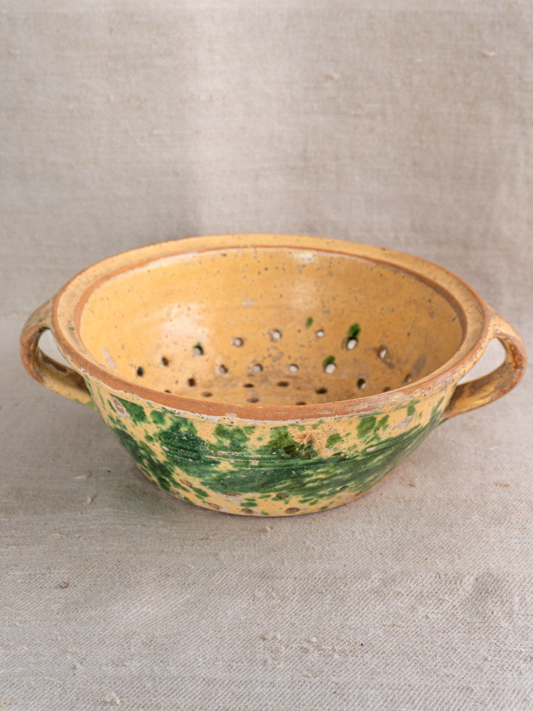 Antique French earthenware colander with jaspe glaze, rustic yellow and green, two handles, shown on neutral fabric.