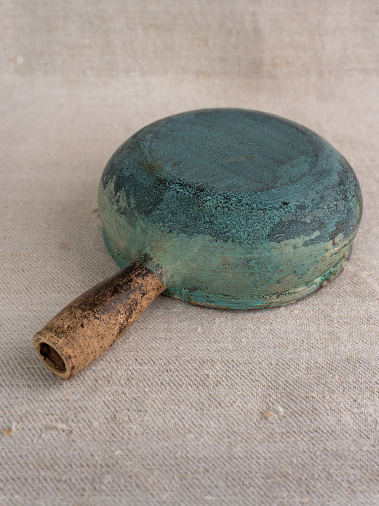 Antique French glazed earthenware poêlon with greenish-blue verdigris finish and wooden handle on beige fabric.