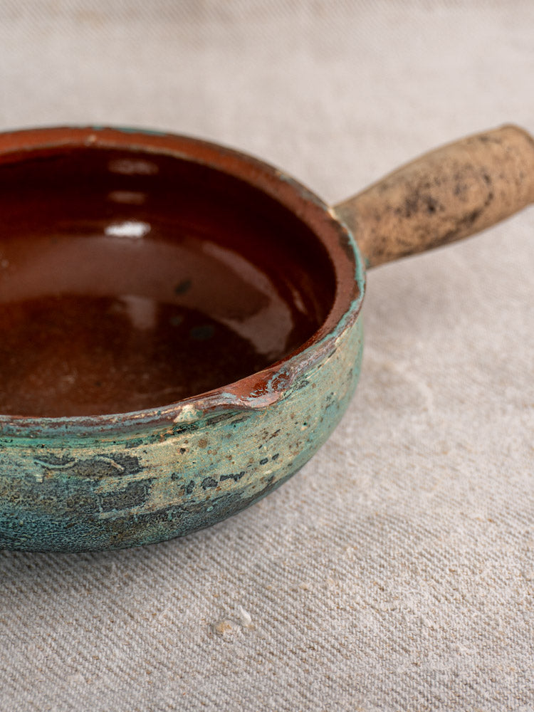 Antique French poêlon glazed earthenware pot with verdigris finish, brown interior, textured handle, on light fabric.