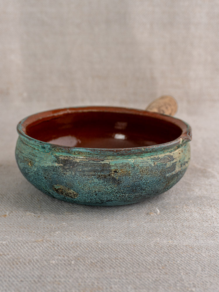 Antique French glazed earthenware poêlon with mottled turquoise and brown glaze, reddish interior, on beige textured fabric.