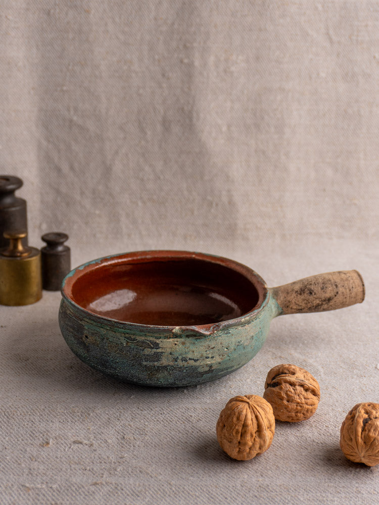 Antique French poêlon glazed earthenware cooking pot with handle on textured fabric, walnuts and metal weights nearby.