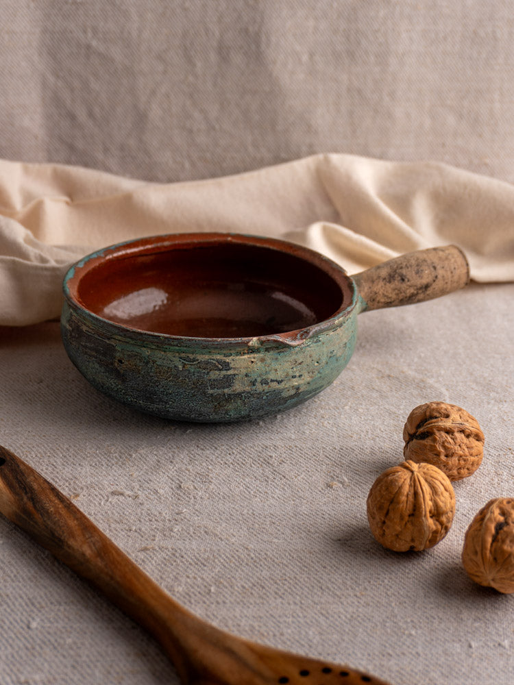 Antique French glazed earthenware poêlon with handle, displayed on textured fabric with walnuts and wooden slotted spoon.