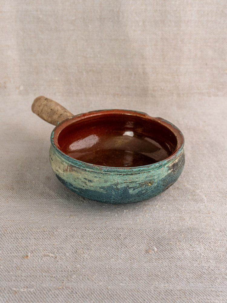 Antique French glazed earthenware poêlon with teal and brown finish, short handle, on textured beige fabric background.
