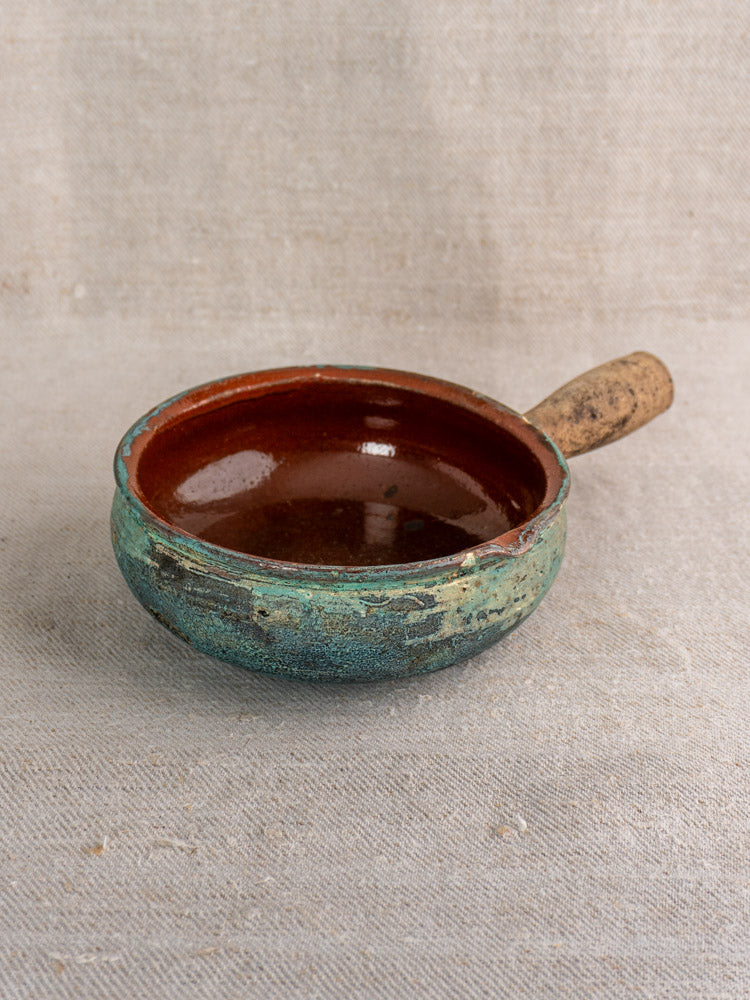 Antique French glazed earthenware poêlon with turquoise verdigris and brown interior, short handle, on beige fabric.