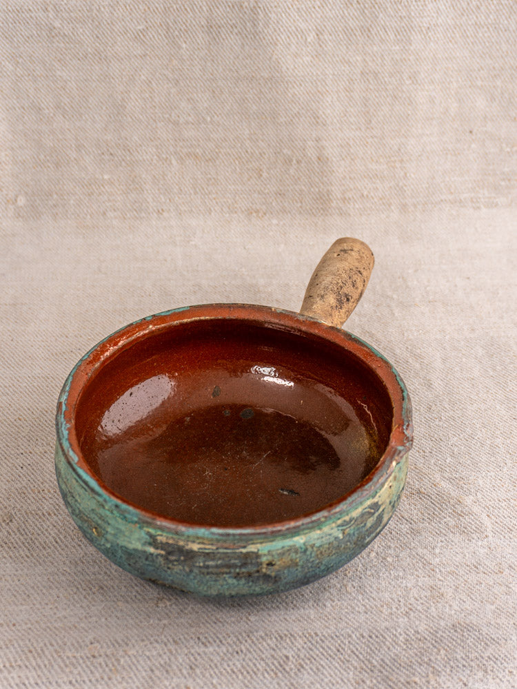Antique French poêlon, round glazed earthenware pot with turquoise verdigris finish, brown interior on beige fabric.