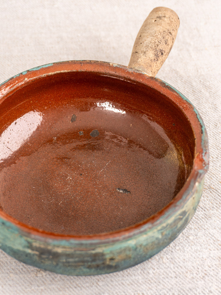 Antique French poêlon, glazed brown earthenware pot with beige handle and worn patina, displayed on light fabric.