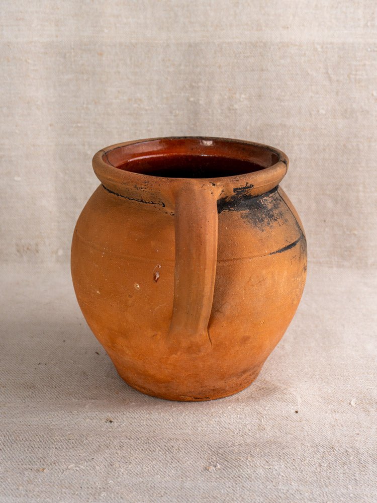 Antique French terracotta cruche with fire-blackened patina and handle, on beige fabric surface.