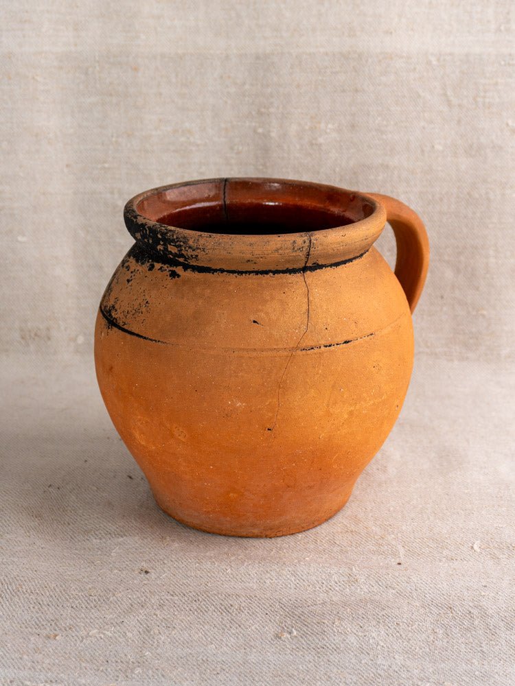 Round orange-brown French terracotta cruche with handle, fire-blackened rim, on beige fabric.