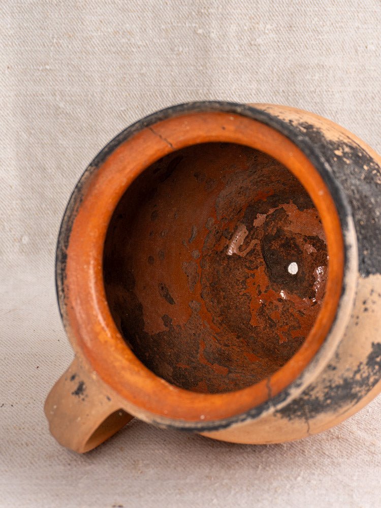 Antique French terracotta mug with fire-blackened patina, single handle, on textured beige fabric.