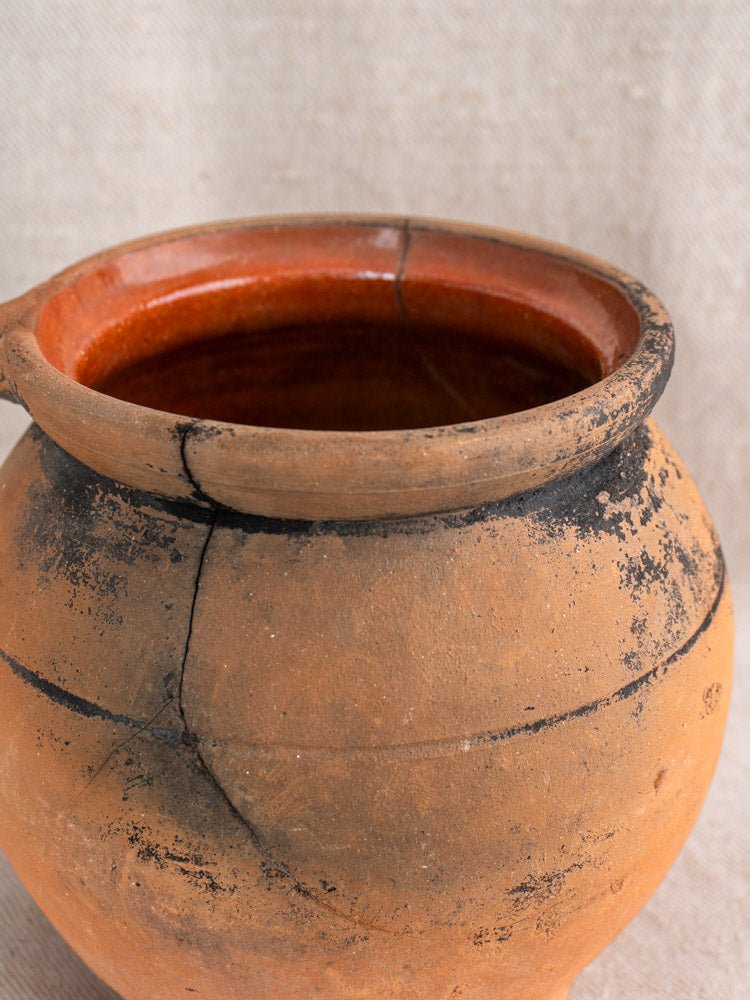 Antique French fire-blackened terracotta cruche with soot marks and cracks on beige fabric background.