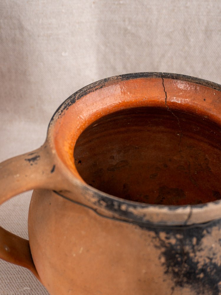 Antique French terracotta cruche pair with cracked, fire-blackened surfaces and uneven rims on beige fabric.