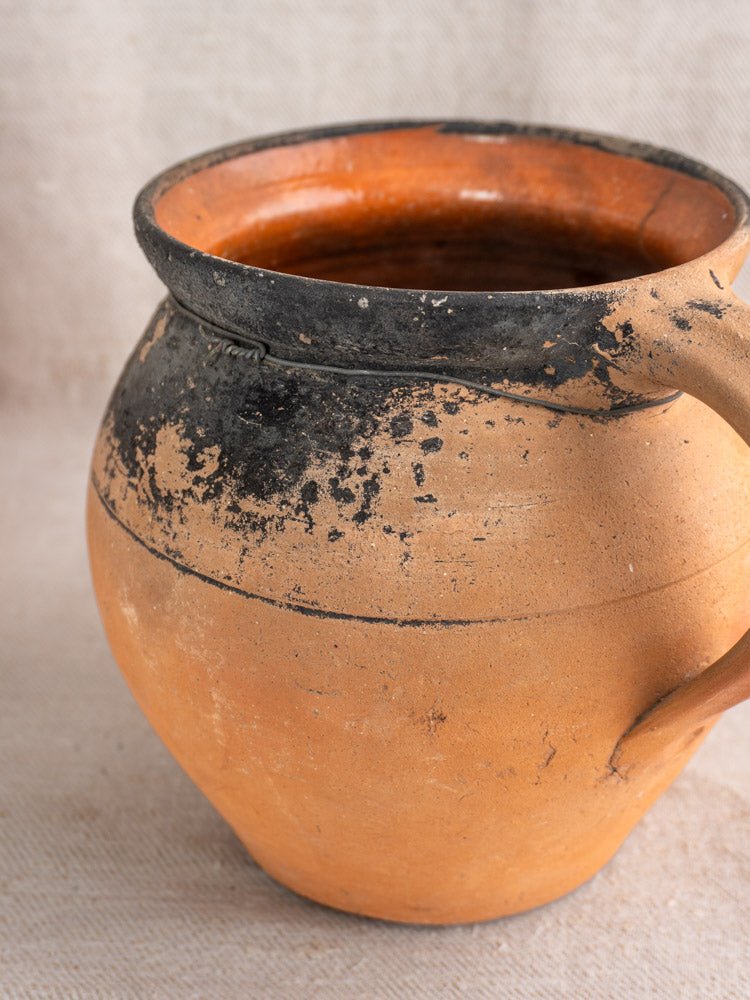 Antique French terracotta cruche with black soot marks, orange-brown patina, on neutral fabric background.