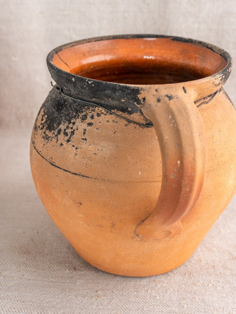 Antique French terracotta cruche with fire-blackened rim, rustic orange clay, single handle, on neutral fabric.