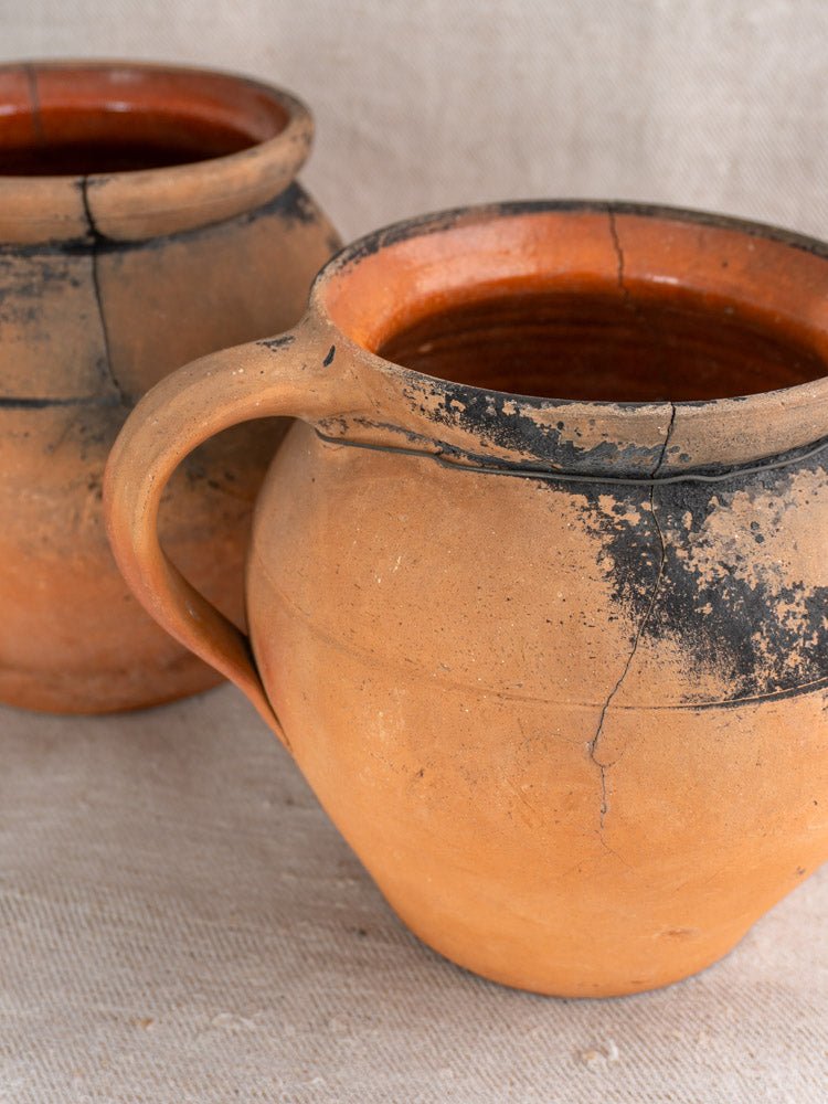 Pair of antique French terracotta cruche water jugs with blackened cracks on beige textured surface.