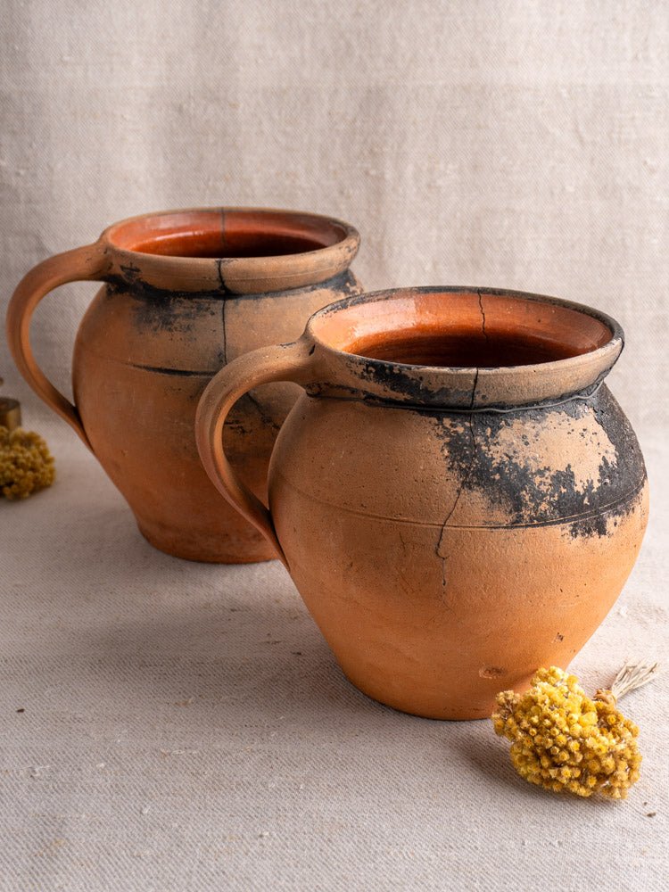 Pair of antique French fire-blackened cruche terracotta jugs with dark burn marks on beige fabric, dried yellow flowers nearby.
