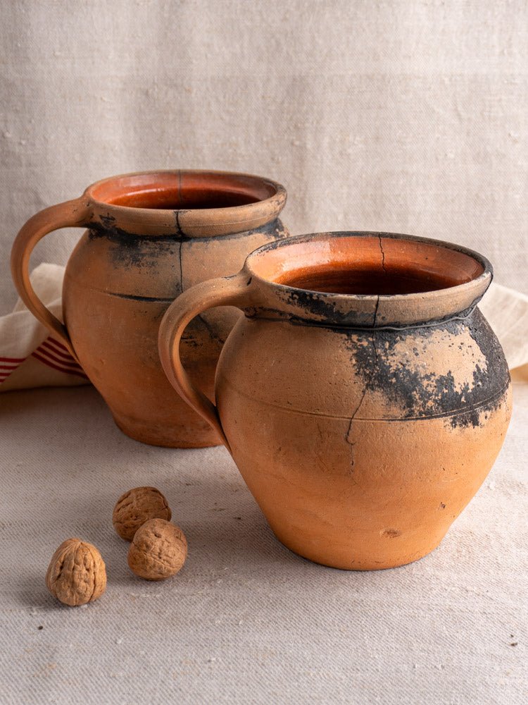 Antique French fire-blackened terracotta cruche water jugs with rustic handles on a neutral fabric, three walnuts nearby.