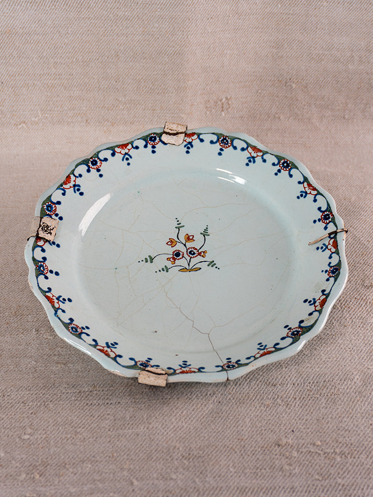 Antique French faience plate with scalloped edges, polychrome floral motifs, staple repairs on neutral textured fabric.