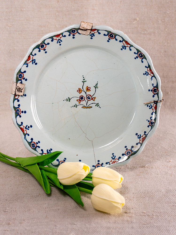 Antique French faience plate with polychrome florals, metal clips; pale yellow tulips on beige fabric backdrop.